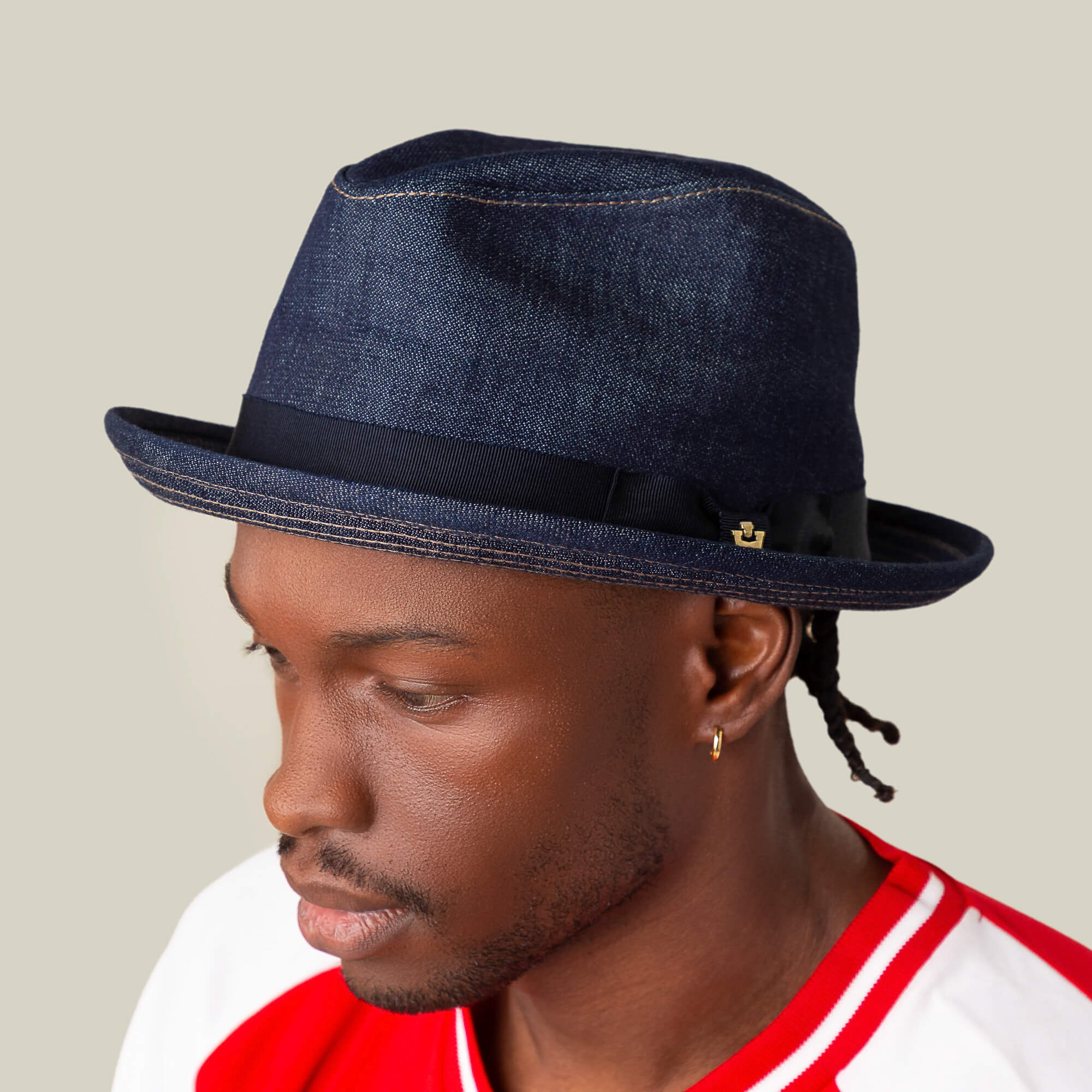 A man wearing the Goorin Bros. Noisy Big Stepper crown fedora in dark blue denim with a black band and pin, paired with a red and white shirt and a small hoop earring.