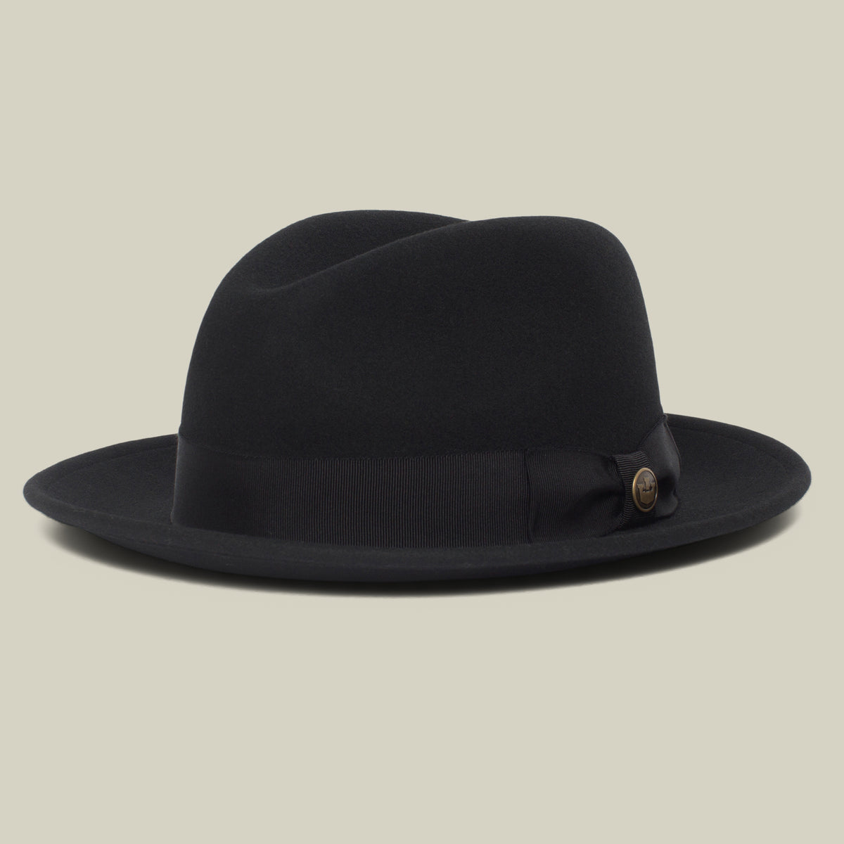 Dean the Butcher by Goorin Bros. is a black fedora hat featuring a slightly curved brim, a black ribbon band, and a small round button on the side. Displayed on a plain beige background, this elegant all-season hat embodies classic fedora style.