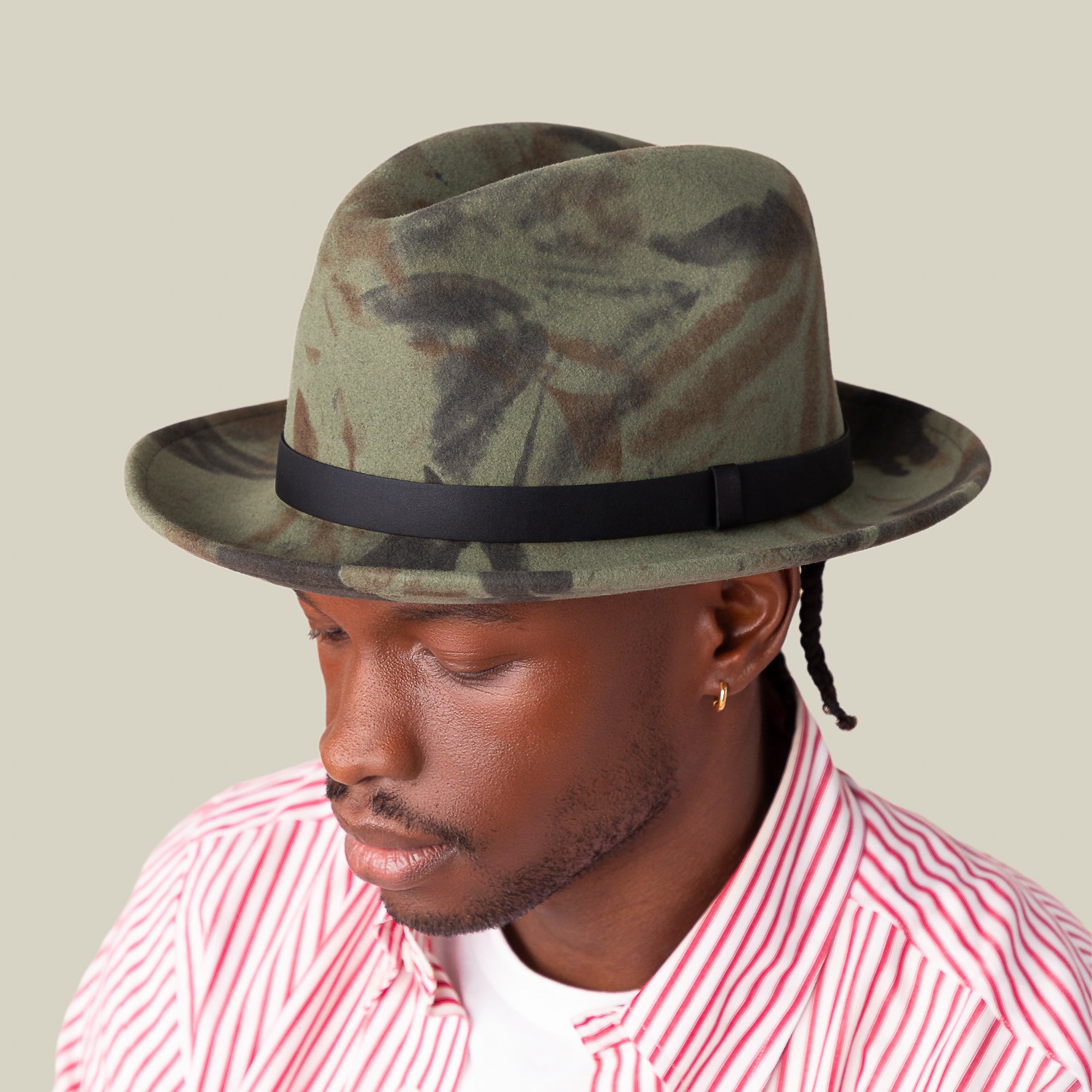 A person wearing the Goorin Bros. "Dean the Butcher" fedora with a green camo pattern and grosgrain hatband, paired with a red-and-white striped shirt and white tee, looks downward against a plain background.