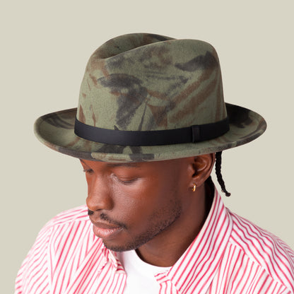 A person wearing the Goorin Bros. "Dean the Butcher" fedora with a green camo pattern and grosgrain hatband, paired with a red-and-white striped shirt and white tee, looks downward against a plain background.