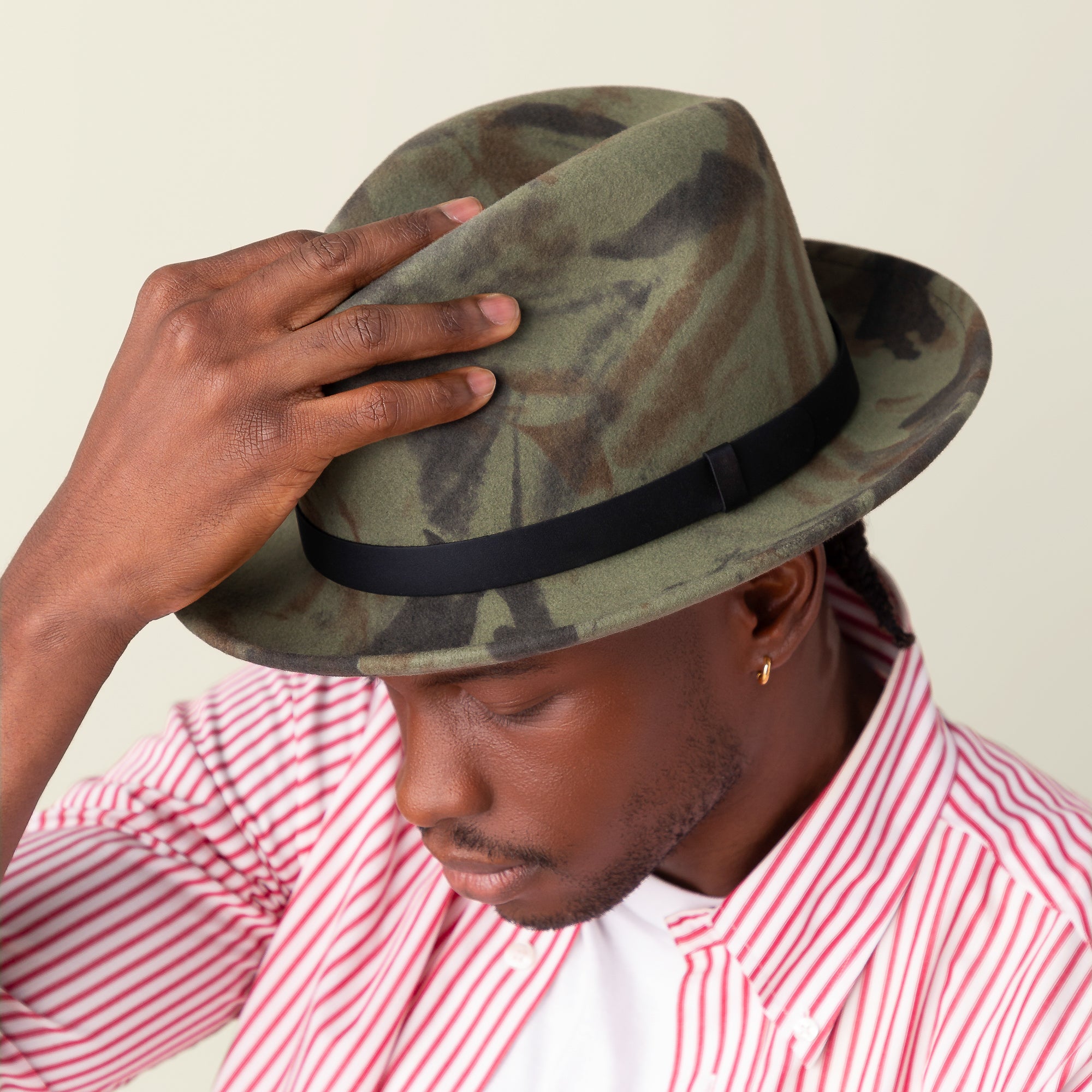 A man tilts his Dean the Butcher fedora by Goorin Bros., featuring green camouflage, while wearing a red and white striped shirt and looking downward.