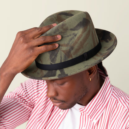A man tilts his Dean the Butcher fedora by Goorin Bros., featuring green camouflage, while wearing a red and white striped shirt and looking downward.