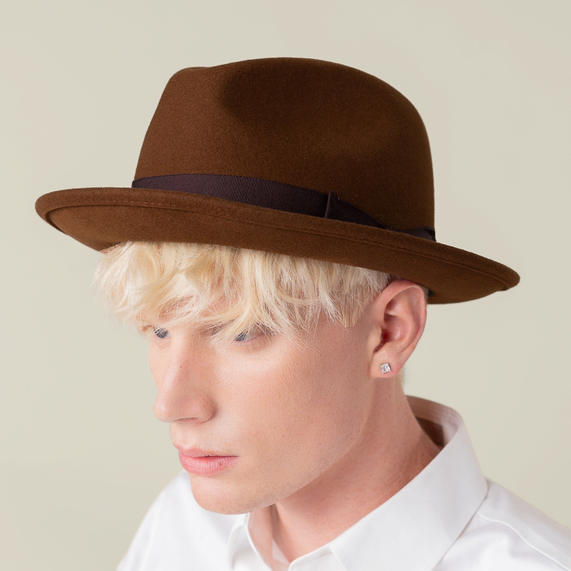 A person with light skin and blond hair wears the Goorin Bros. "Dean the Butcher" brown fedora, a white collared shirt, and a stud earring, facing slightly left against a neutral background.