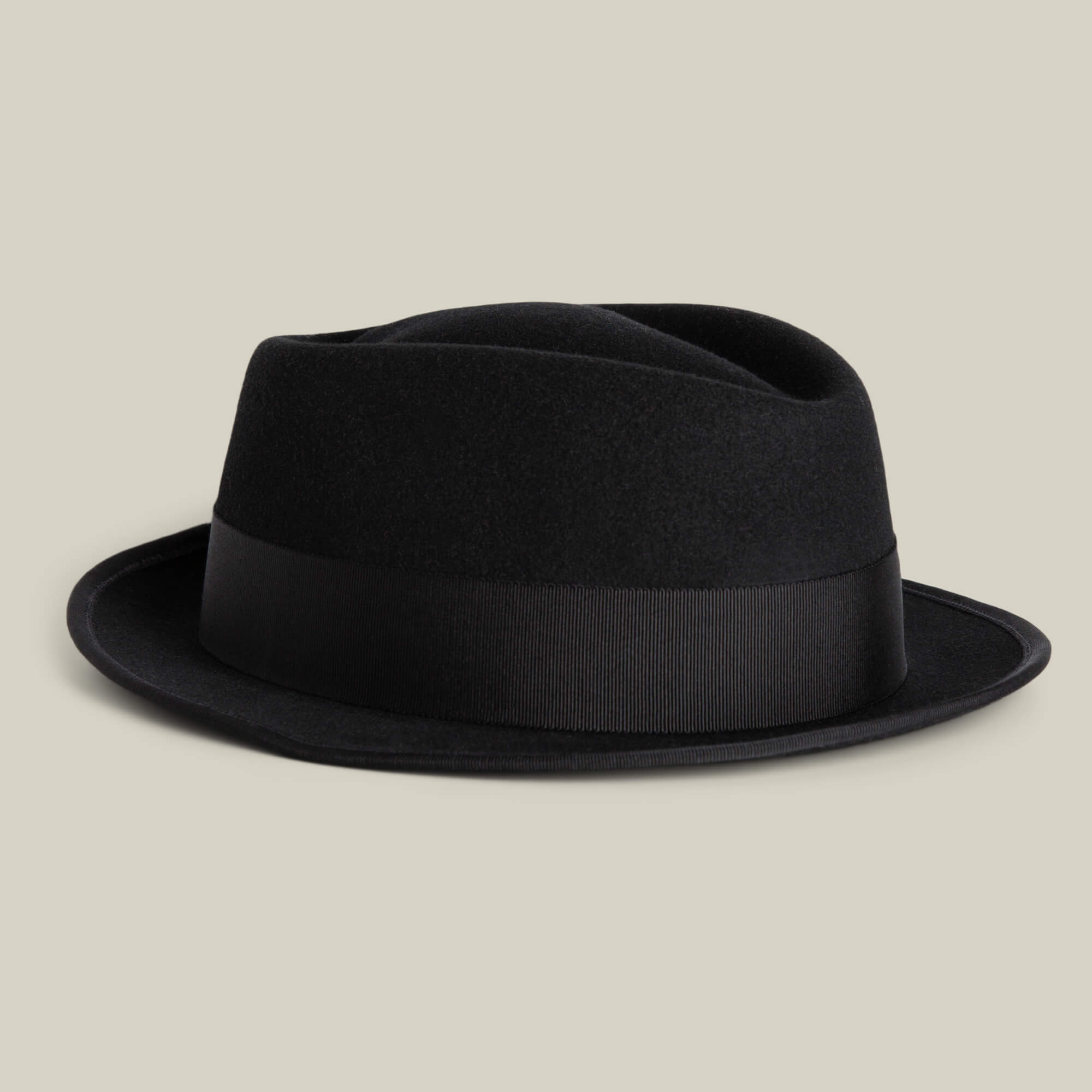 The Slick Carter by Goorin Bros. is a black wool fedora with a short brim and wide black ribbon band, shown on a light beige background for effortless style.