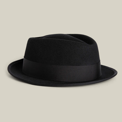 The Slick Carter by Goorin Bros. is a black wool fedora with a short brim and wide black ribbon band, shown on a light beige background for effortless style.