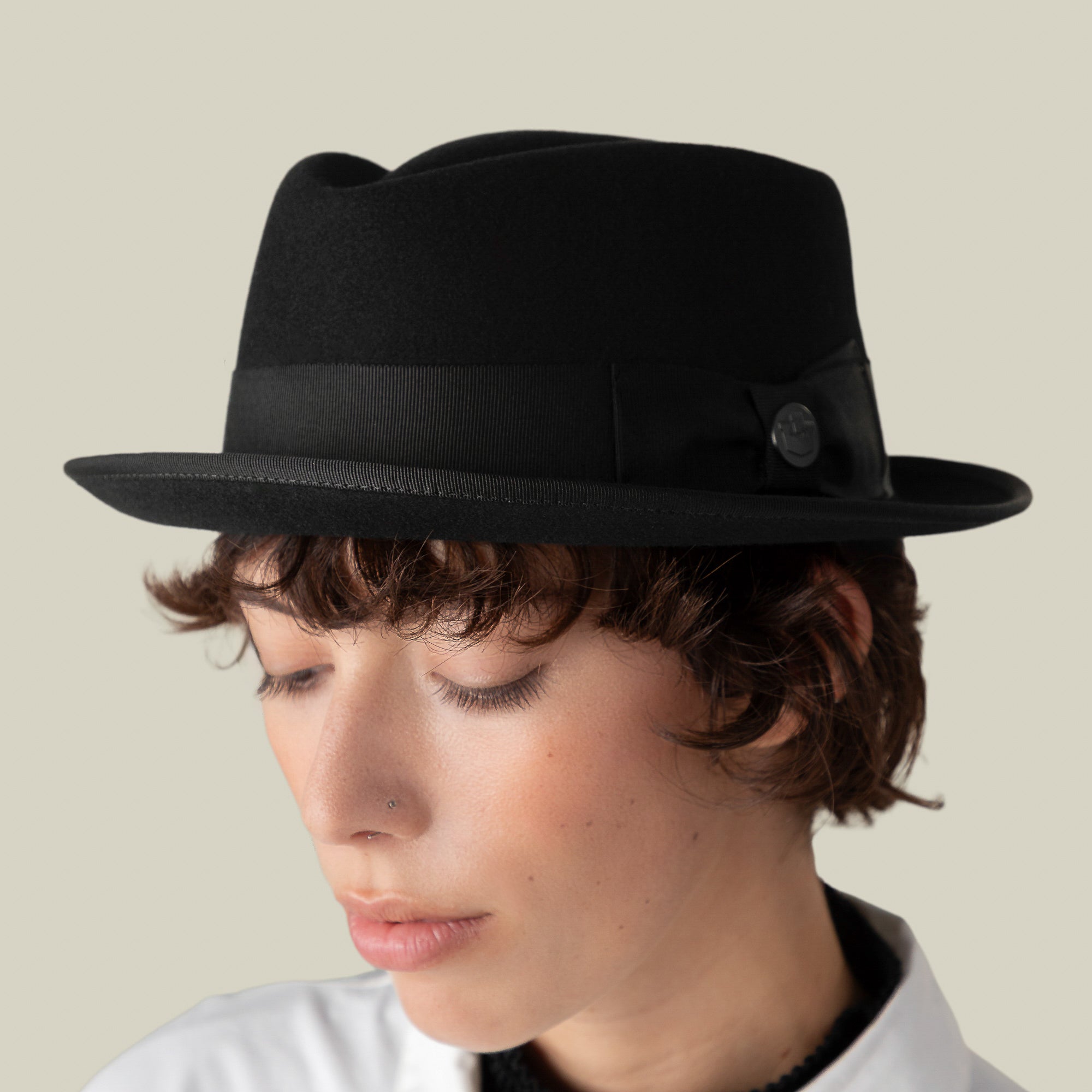 A person with short brown hair wears the Goorin Bros. Slick Carter black wool short-brim fedora and a white collared shirt, looking downward against a plain light background, capturing the brand's effortless style.
