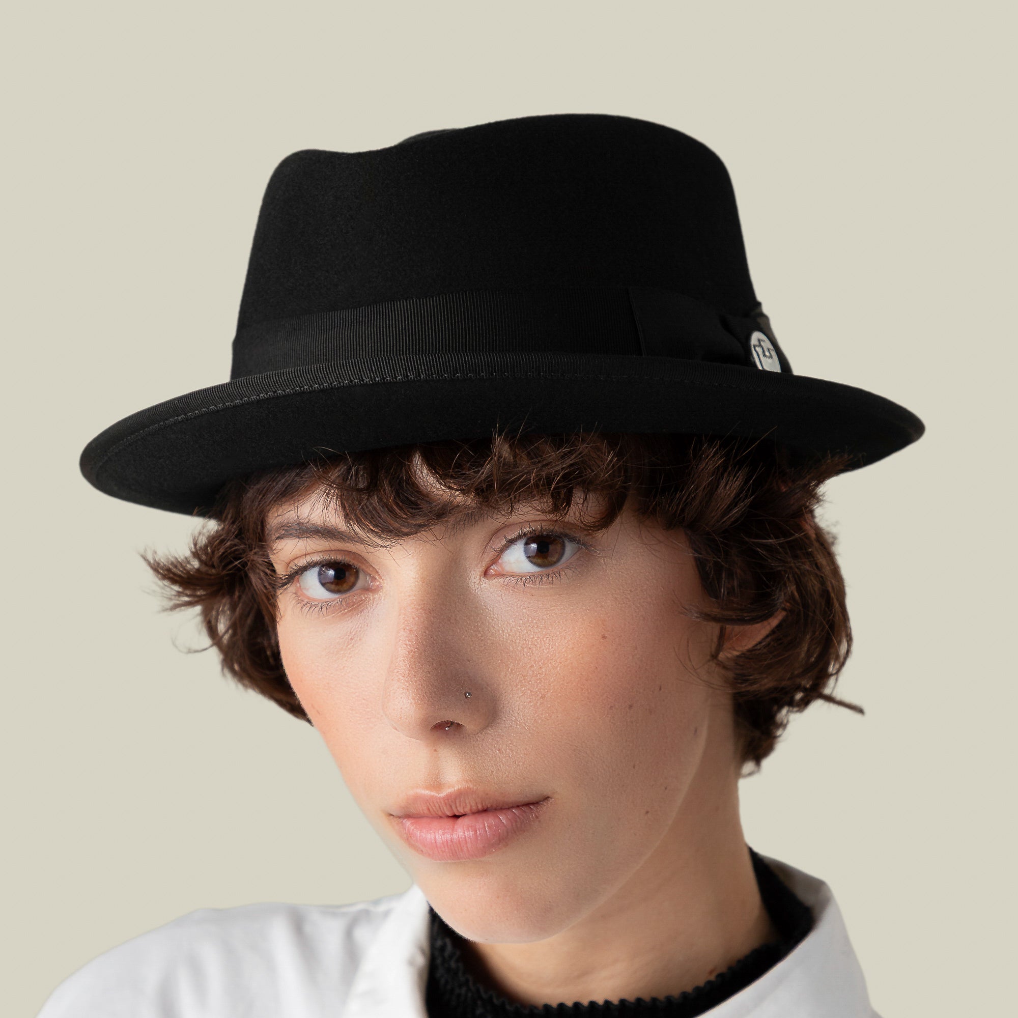 Person with short curly hair wearing the Slick Carter wool short brim black fedora by Goorin Bros. and a white shirt, looking directly at the camera against a plain background, radiating effortless style.