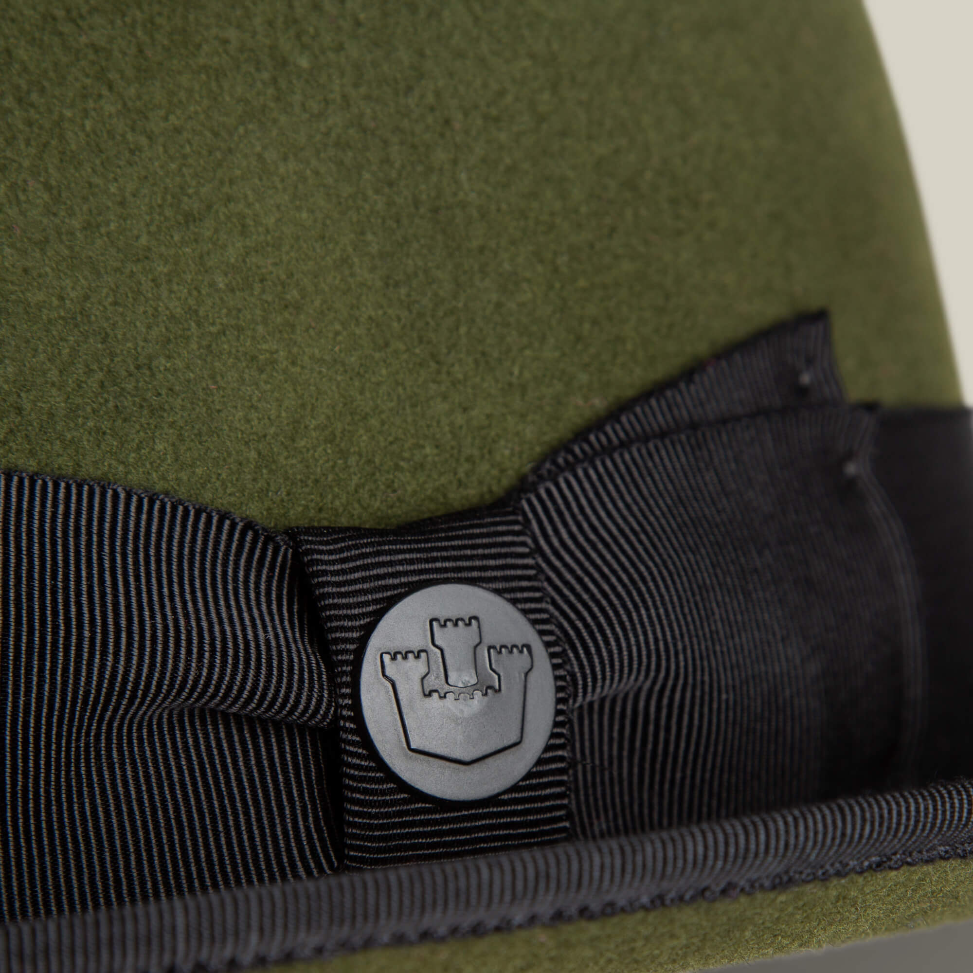 Close-up of an olive green wool short brim felt hat by Goorin Bros., featuring a black grosgrain ribbon band and round metal emblem with a castle icon—effortless Slick Carter style.
