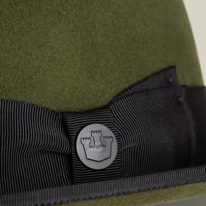 Close-up of an olive green wool short brim felt hat by Goorin Bros., featuring a black grosgrain ribbon band and round metal emblem with a castle icon—effortless Slick Carter style.