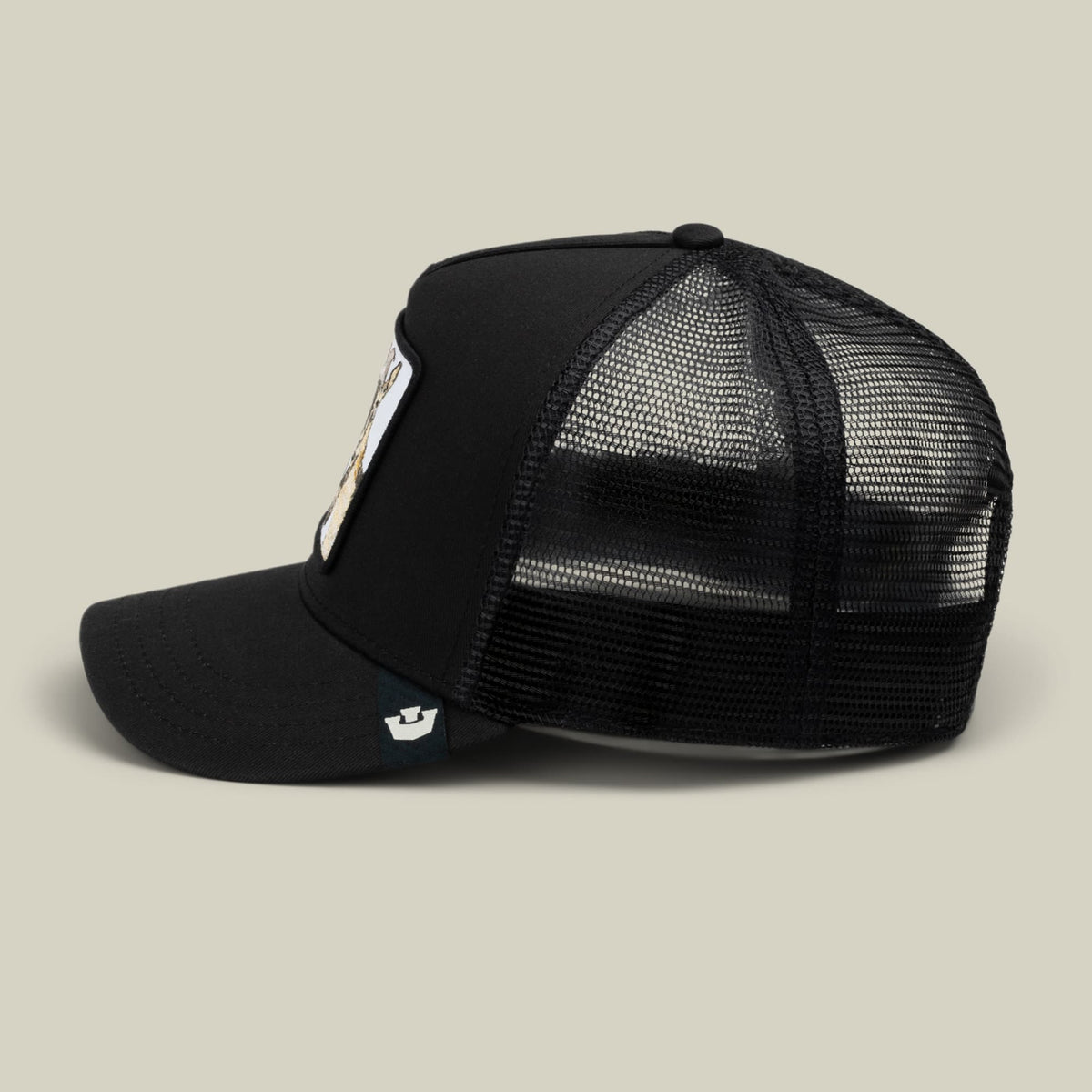 The Goorin Bros. Goat Beard black trucker hat features a mesh back panel, snapback closure, and a small embroidered patch on the front, displayed against a neutral beige studio background.