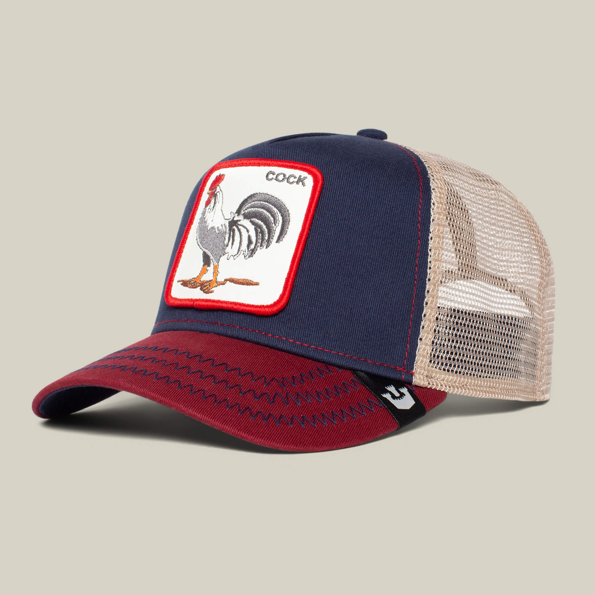 The Farm’s "The Cock" trucker hat has a navy blue and red front, beige mesh back, and a patch with a rooster illustration and the word COCK for a bold statement.