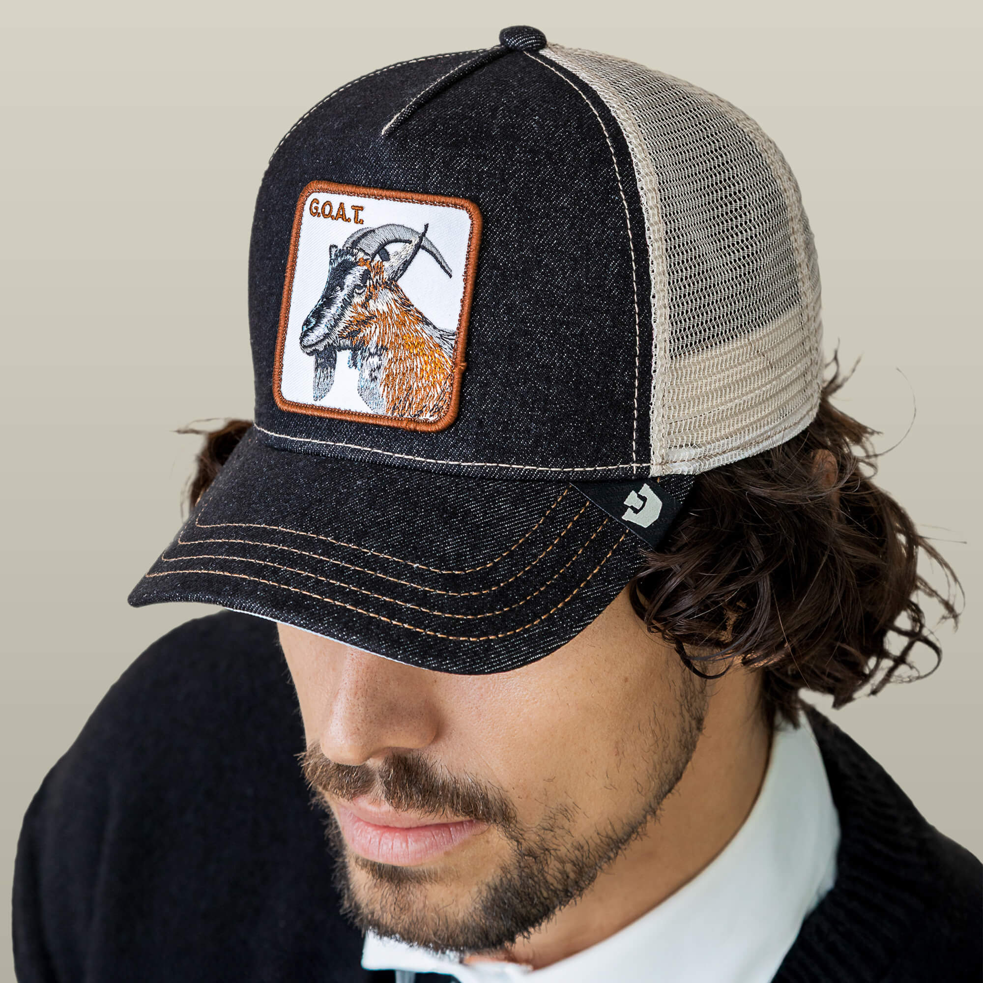A man wears The Farm's "The GOAT" hat in black and beige mesh, featuring a bold front patch with a goat and "GOAT"—the perfect pick for anyone seeking the Greatest Of All Time hat.