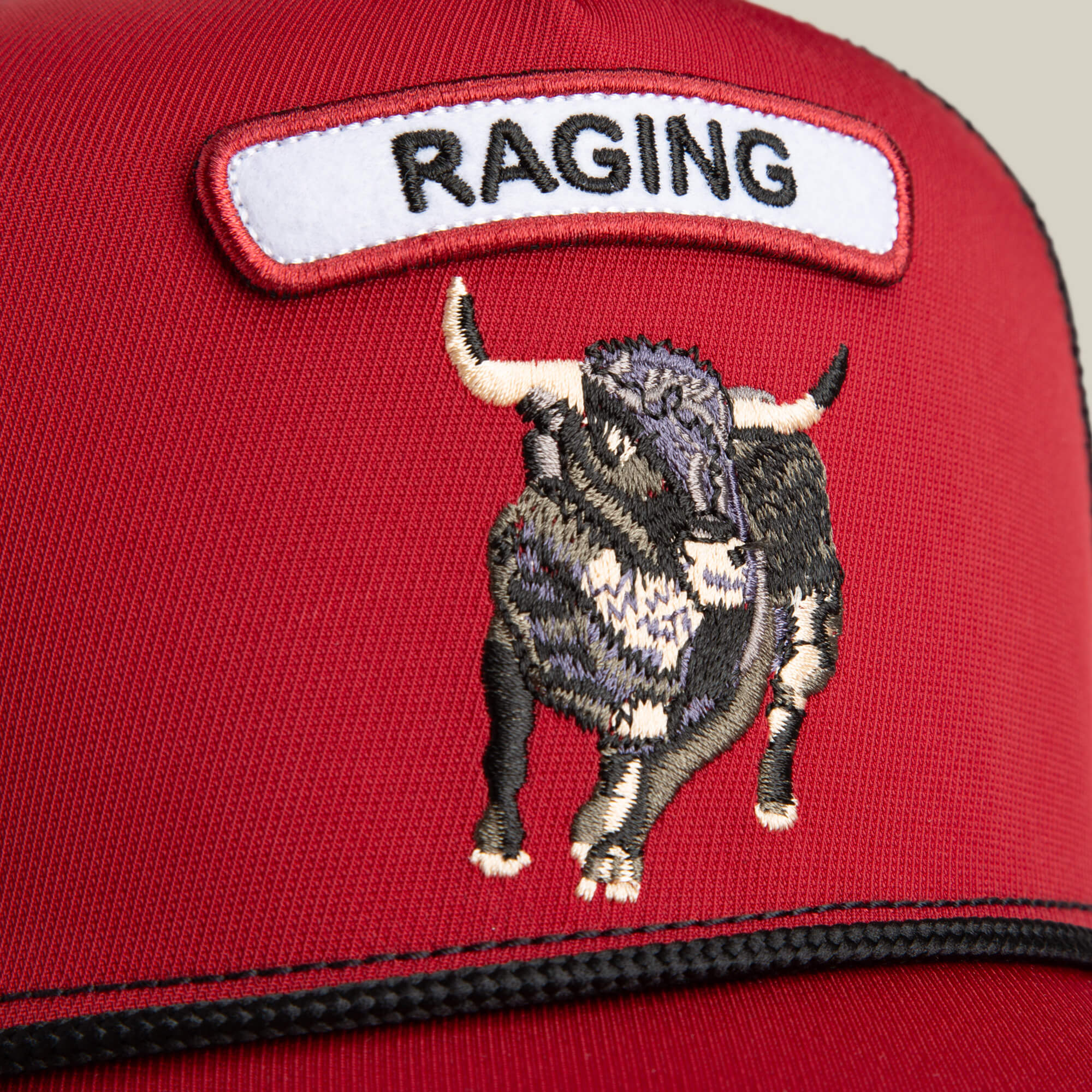 Close-up of the Goorin Bros. GB2 Raging Bull trucker cap with a red mesh crown, embroidered bull, and a white patch featuring “RAGING” in black letters.