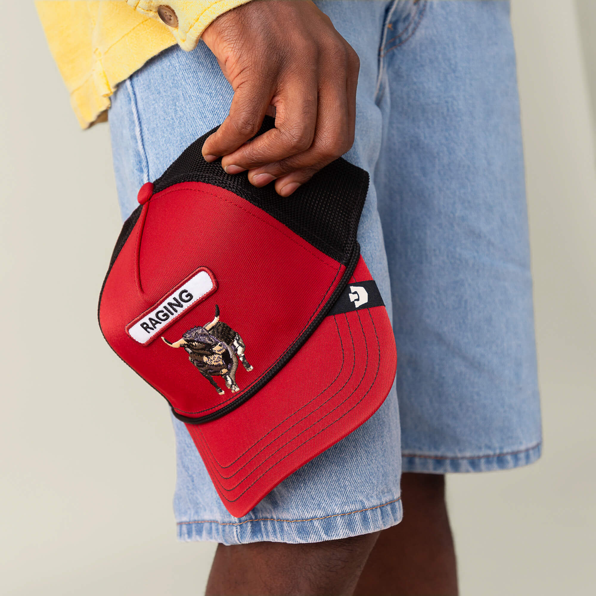 Wearing denim shorts and a yellow shirt, a person holds the Goorin Bros. GB2 Raging Bull red crown hat, featuring a bull patch and the word "RAGING" on the front.