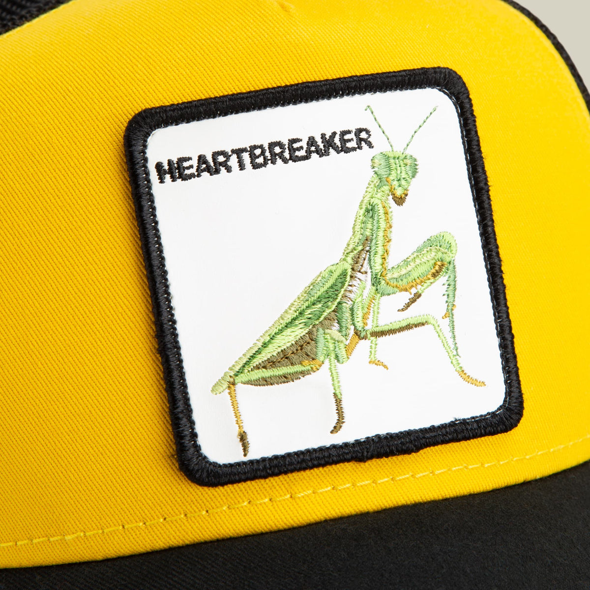 The Heartbreaker by Goorin Bros is a yellow and black trucker hat featuring an embroidered praying mantis patch on the front. The close-up studio shot highlights the detailed insect motif and classic baseball cap silhouette.