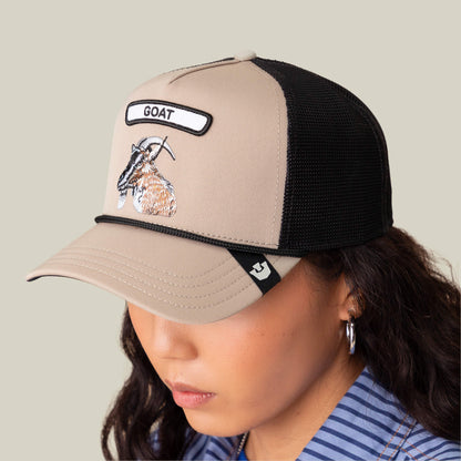 Someone wears The Farm’s GB2 Goat Trucker Hat in beige and black, featuring an embroidered goat and “GOAT” on the front panel—a true badge of honor.