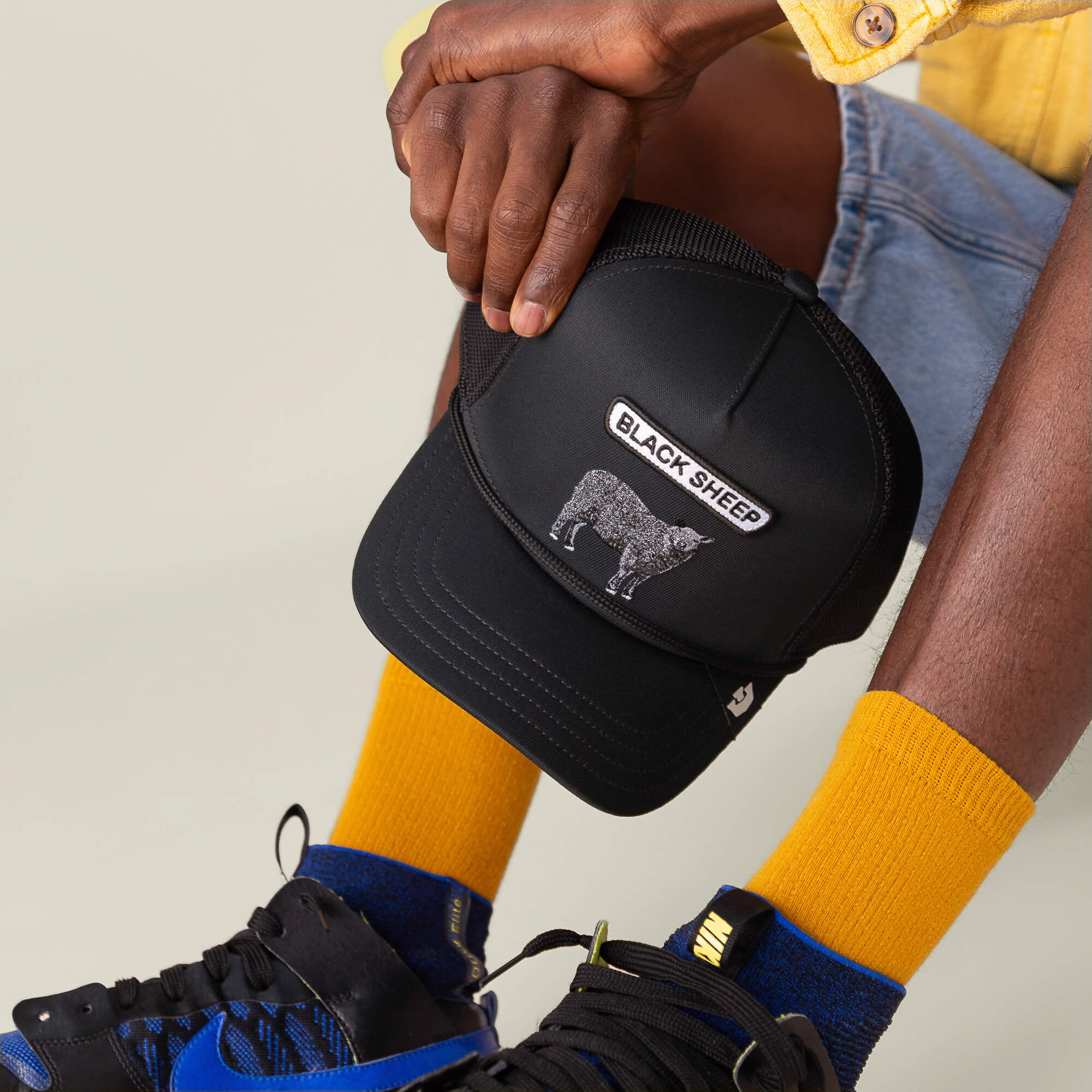 A person in yellow socks and blue-black sneakers holds The Farm's GB2 Black Sheep cap, its patch as distinctive as a finely tuned engine.