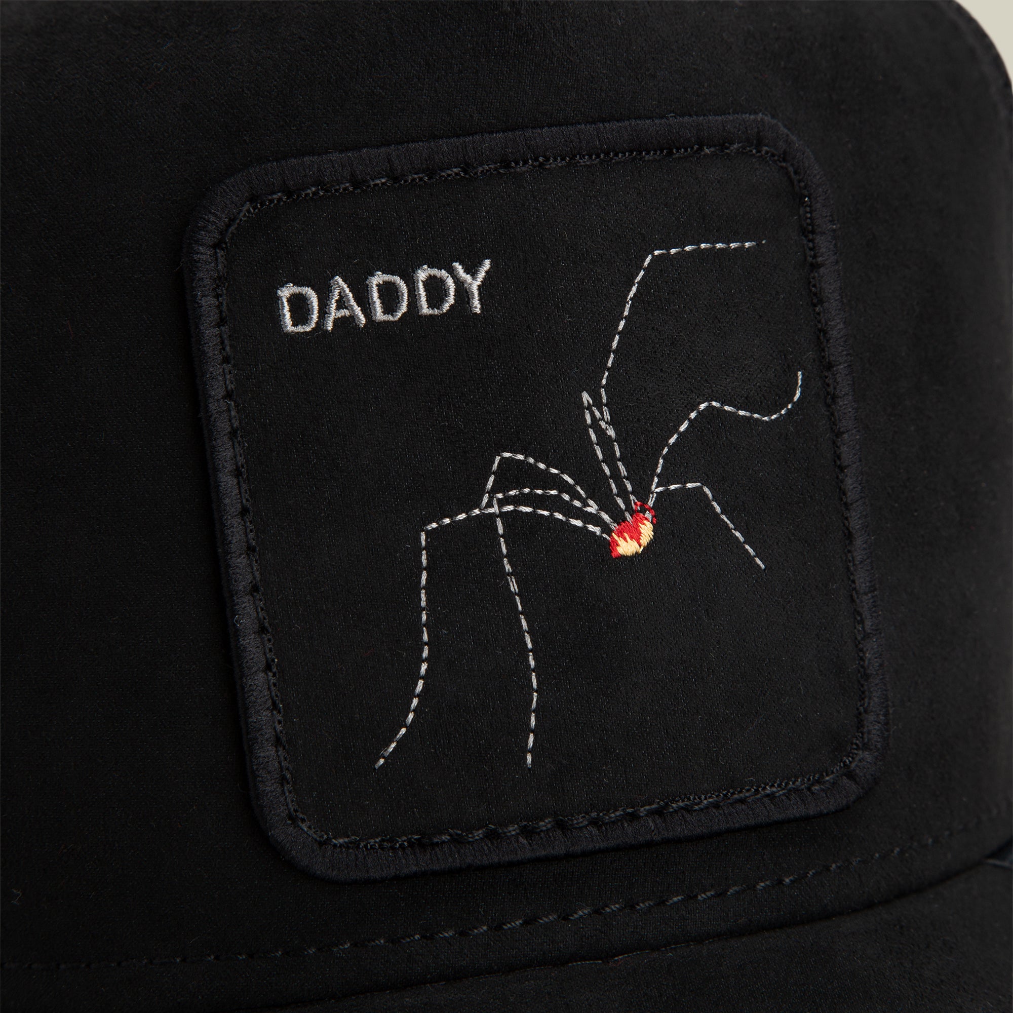 A black fabric patch from Goorin Bros., named The Suede Spider, featuring the word “DADDY” and an embroidered arachnid with a red spot, is sewn onto a black surface.