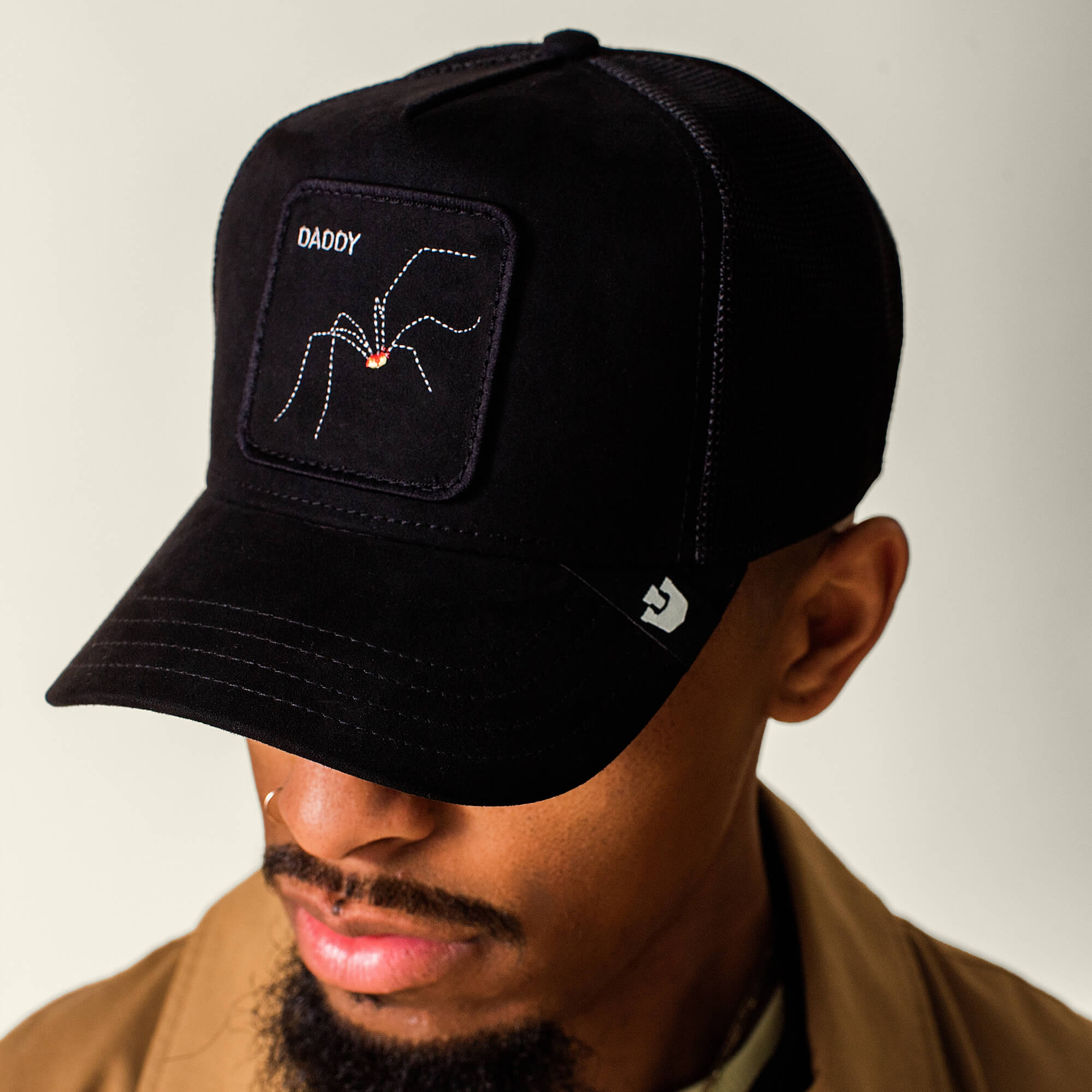 A man wearing The Suede Spider hat by Goorin Bros.—a black microsuede cap featuring a spider arachnid and the word “DADDY” embroidered on the front patch—looks downward.