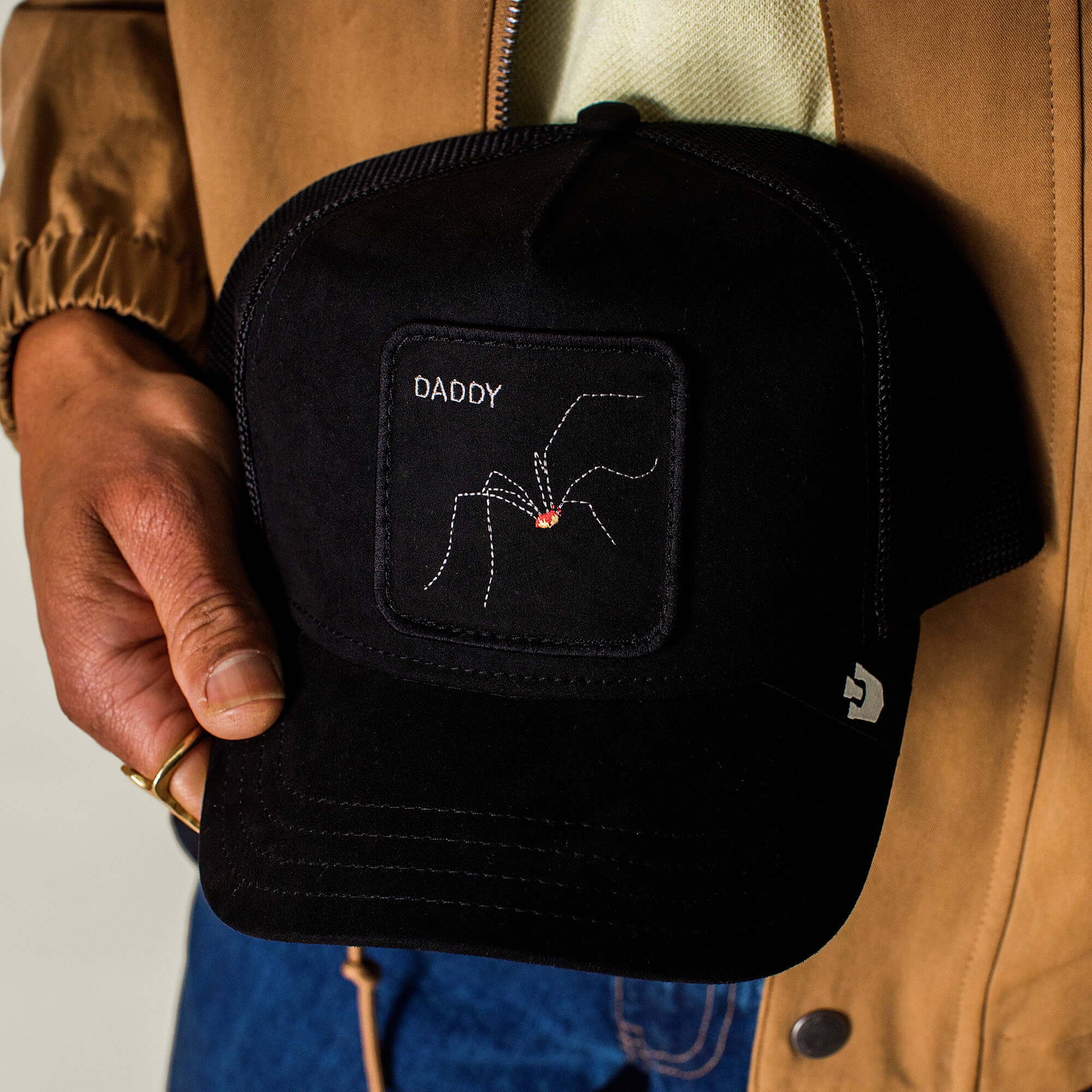 A person holds The Suede Spider by Goorin Bros., a black microsuede trucker cap featuring a patch with an embroidered arachnid and the word “DADDY” above it—perfect for fans of dad jokes and unique headwear.