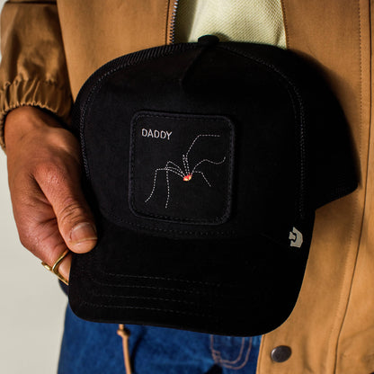 A person holds The Suede Spider by Goorin Bros., a black microsuede trucker cap featuring a patch with an embroidered arachnid and the word “DADDY” above it—perfect for fans of dad jokes and unique headwear.