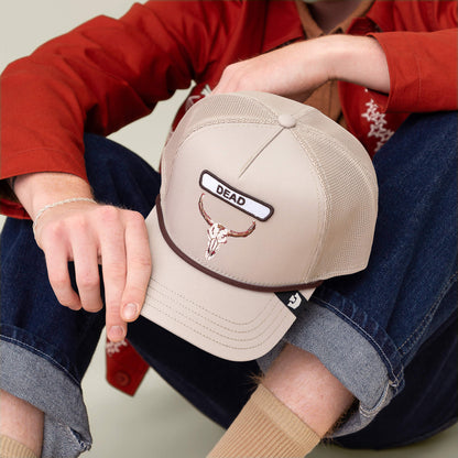 Someone in rolled-up jeans and a red jacket holds the Goorin Bros. GB2 Skull trucker hat, which features a skull patch and "DEAD" embroidered on the front in rugged desert tones.