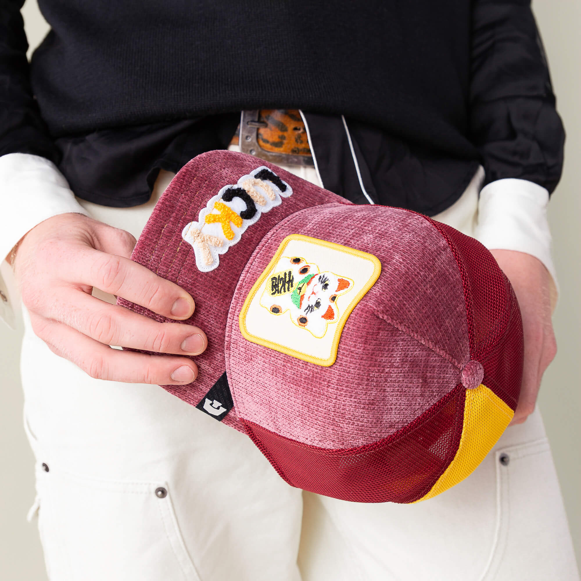 A person in black and white holds a red and yellow Goorin Bros. Maneki-Neko trucker hat with decorative patches, including a playing card shape, while carrying a plush LUCKY beanie under their arm.