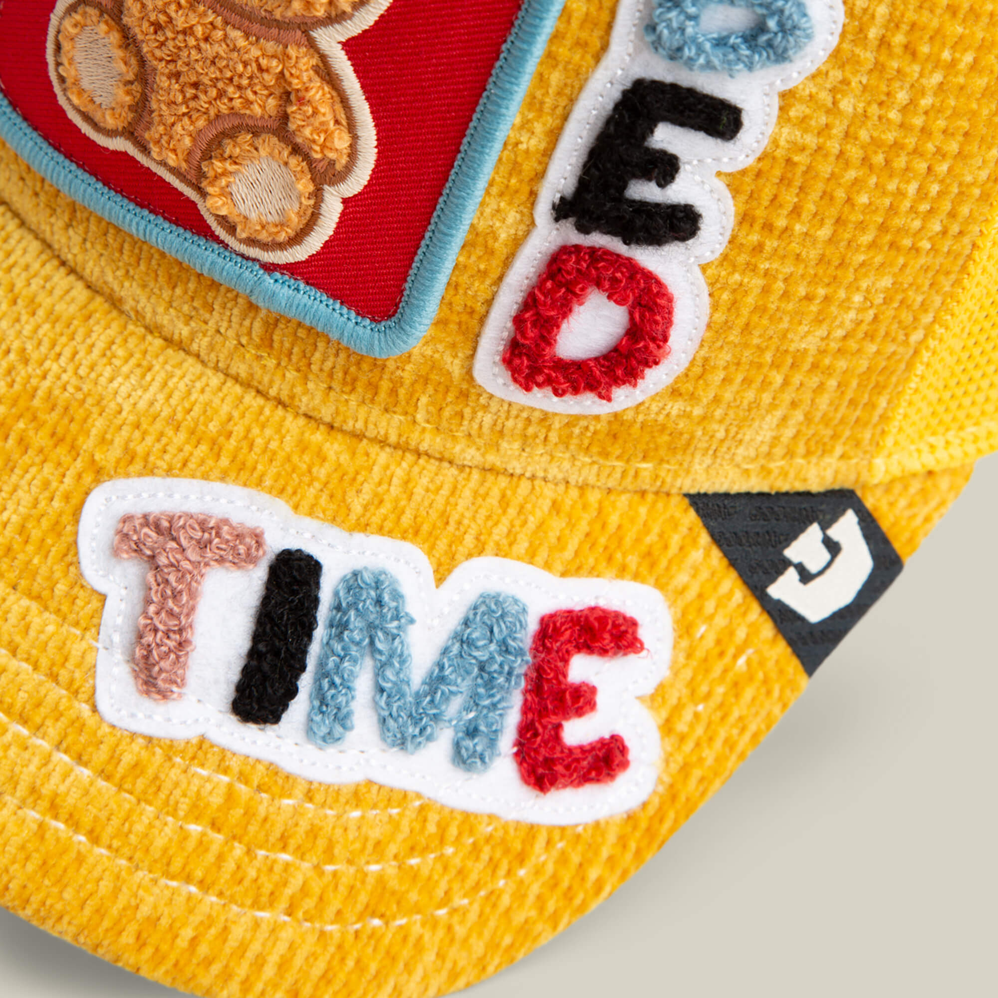 Close-up of the Goorin Bros. Duvet All Det hat in sunshine yellow corduroy with colorful chenille patches spelling "TIME" and part of "BED," plus a small embroidered teddy bear. Ideal for Bed Time Energy style fans.