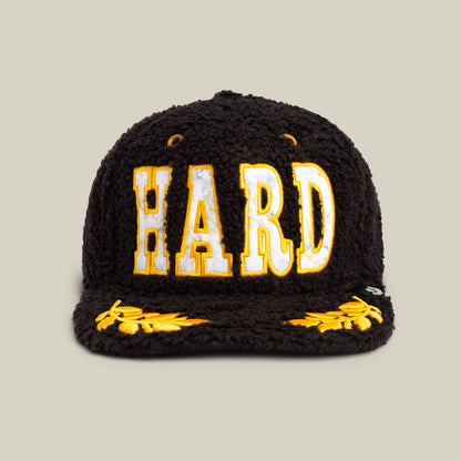 The Goorin Bros. Marble Madness cap features a black textured design, bold yellow and white "HARD" lettering on the front, gold details, and yellow embroidered leaf patterns on the brim.