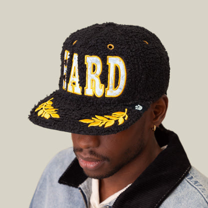 A man wears the Goorin Bros. Marble Madness textured black cap with bold yellow and white HARD lettering, gold accents, and yellow leaf embroidery, styled with a light denim jacket for an amplified silhouette.