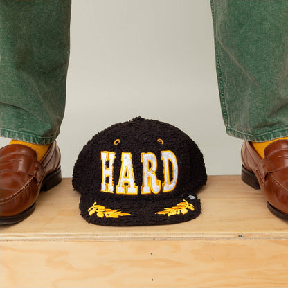 The Goorin Bros. Marble Madness black sherpa-textured cap, featuring "HARD" in bold yellow and white letters, is displayed on a wooden surface between two feet in brown loafers and green pants.