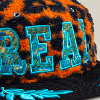 A close-up of the El Camino cap by Goorin Bros. in orange leopard sherpa with blue embroidered letters, a black brim, and an aqua embroidered leaf design.