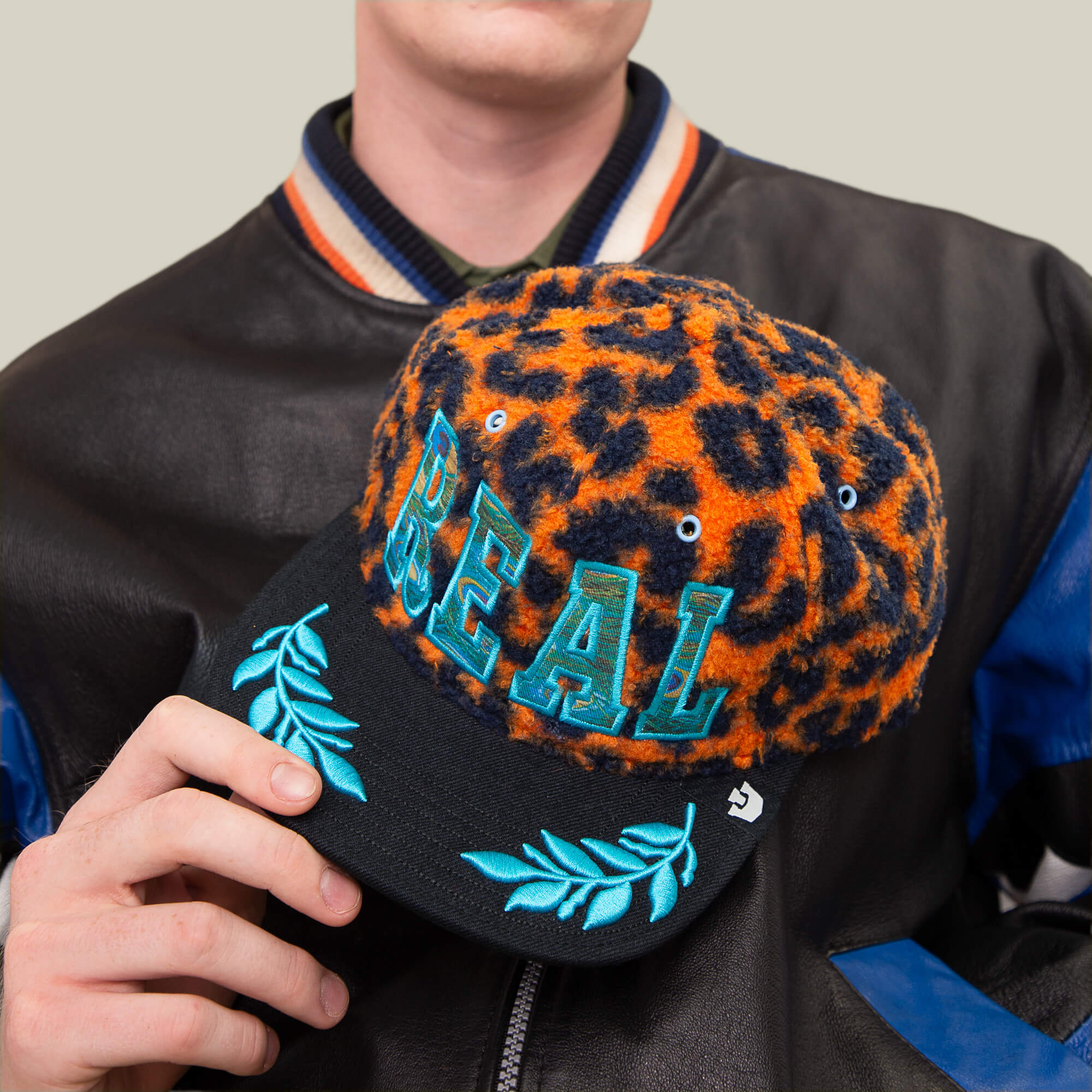 A person holds the Goorin Bros. El Camino cap—an orange leopard sherpa hat with bold blue REAL text and aqua embroidery on the brim—while wearing a black and blue varsity jacket.