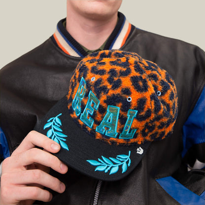 A person holds the Goorin Bros. El Camino cap—an orange leopard sherpa hat with bold blue REAL text and aqua embroidery on the brim—while wearing a black and blue varsity jacket.