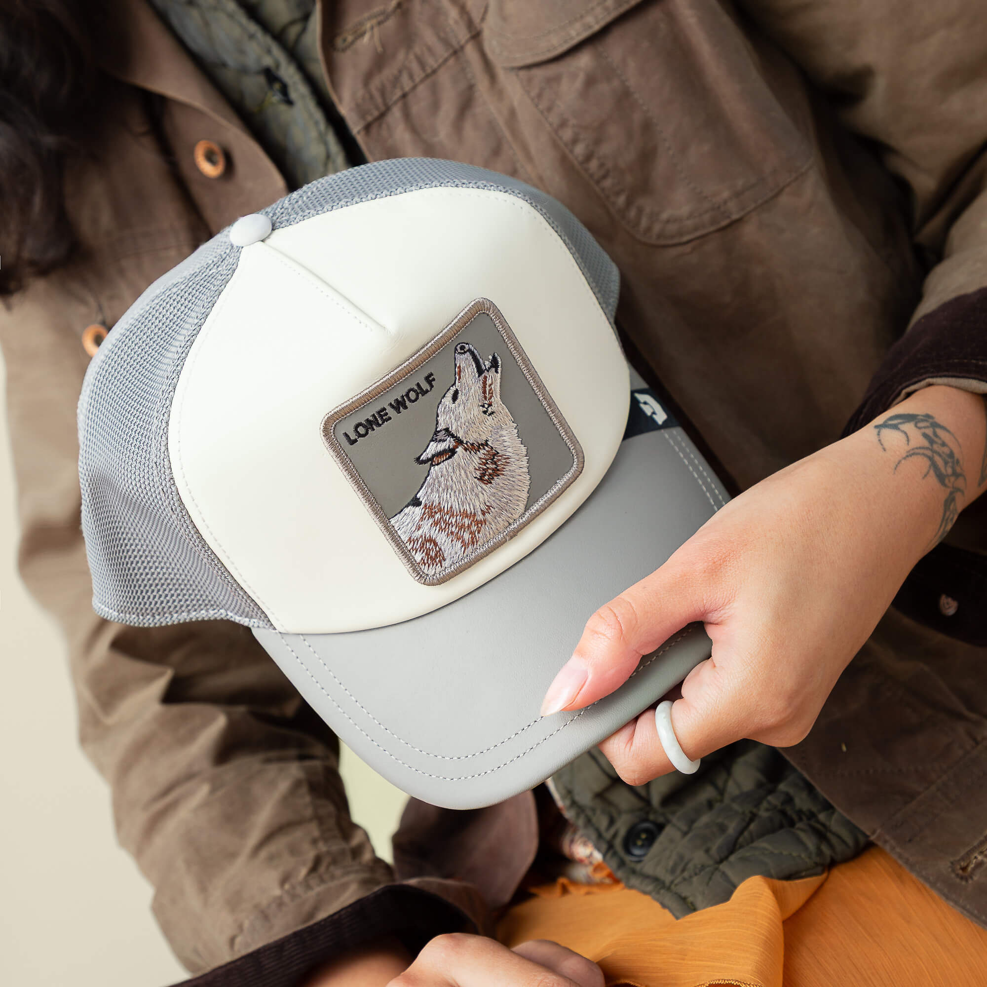 A person holds the Goorin Bros. LFG Lone Wolf gray and white trucker hat with a wolf patch, pairing it effortlessly with courtside sneakers for the ultimate tunnel fit look.
