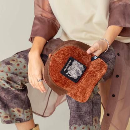 A person holds the Goorin Bros. "Iced Rhino" cap, styled with patterned pants, a sheer brown shirt, a beige top, and velvet rust accents.
