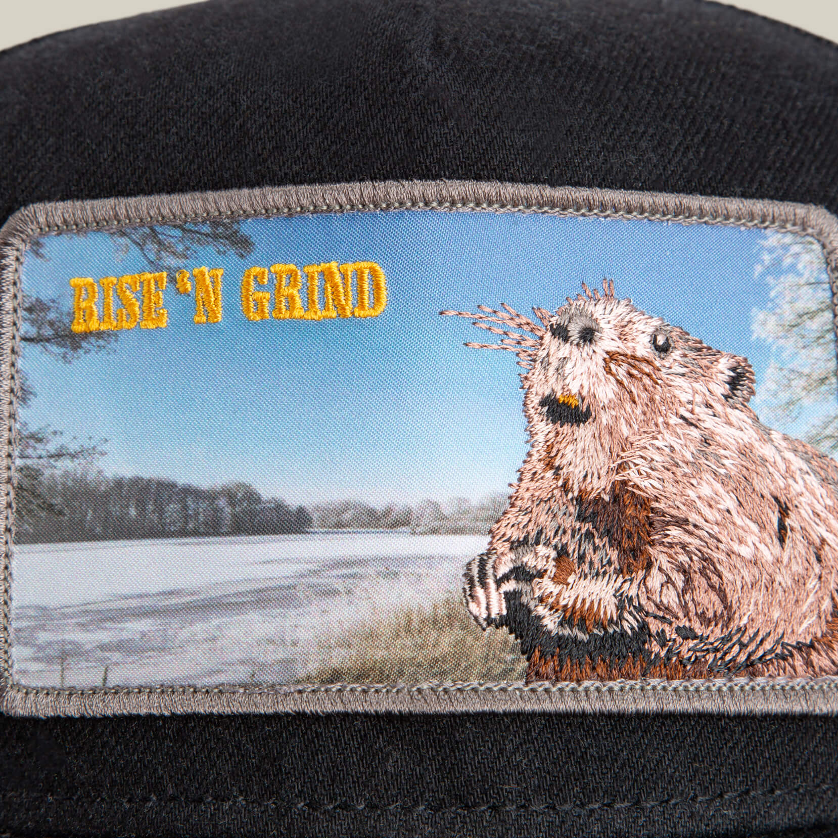 A close-up of the Goorin Bros. Morning Wood cap’s black crown displays an embroidered beaver and the text RISE ‘N GRIND, set against a snowy landscape.