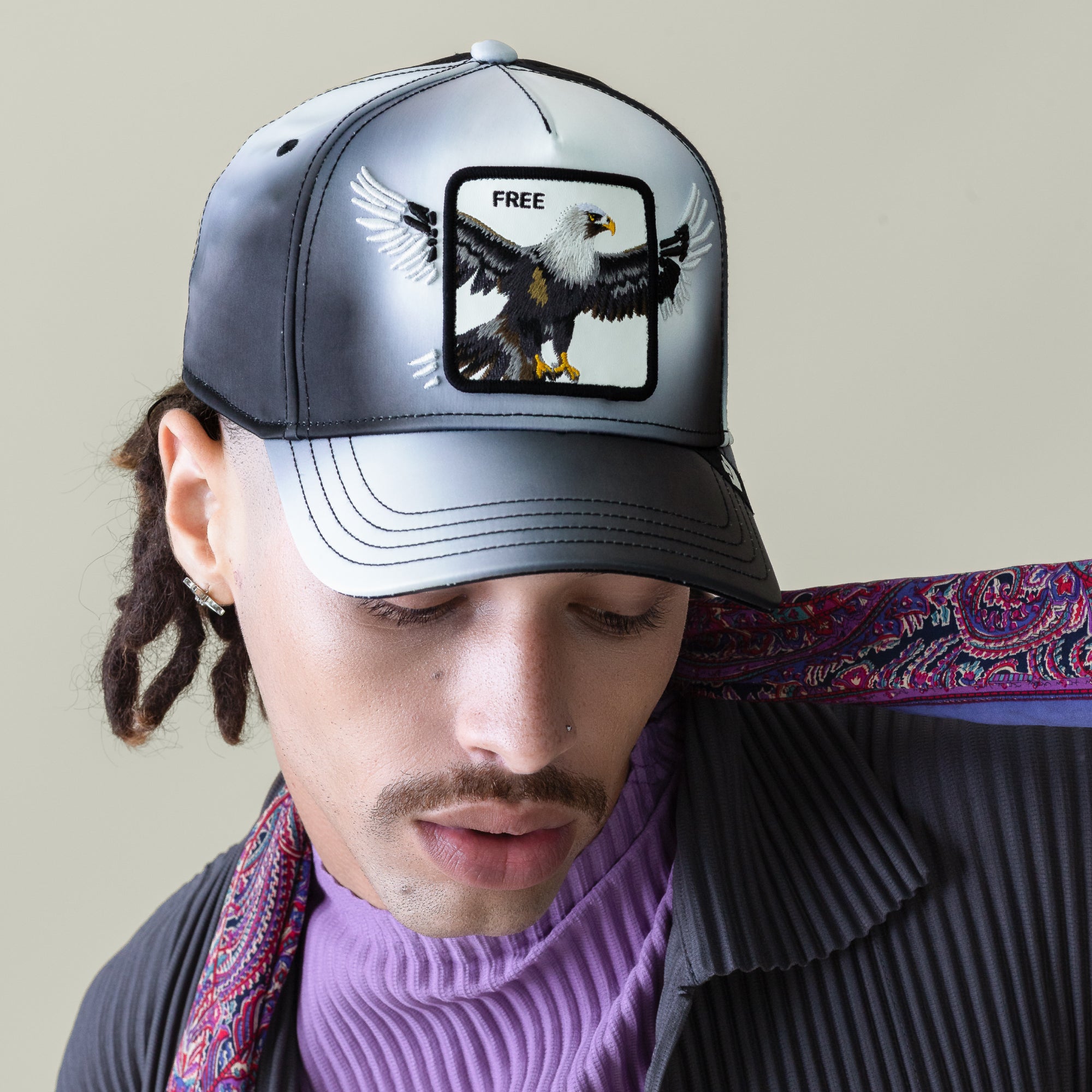 Someone sporting the Goorin Bros. "Steal This Hat," a black-and-grey gradient cap featuring a bold bald eagle patch and "FREE" on the front, paired with a purple turtleneck, pinstriped jacket, and patterned scarf.