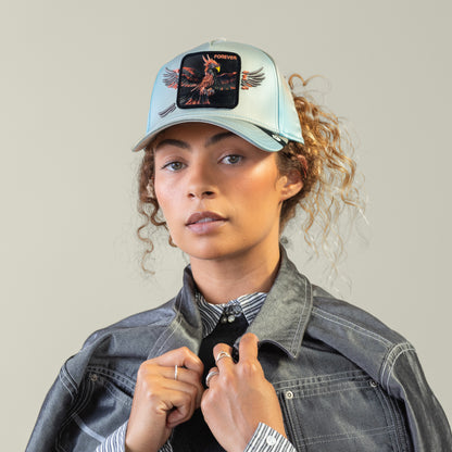 A woman in a sky-blue Goorin Bros. "Never Stop Never Stopping" crown cap with a bird and FOREVER patch, black shirt, and gray denim jacket draped over her shoulders exudes timeless style against a plain background.