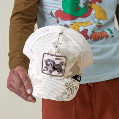 Someone holds the Bestie Camo cap from Goorin Bros., a soft corduroy hat in off-white snow camo with a “BESTIE” shaggy pup patch, paired with a cartoon graphic tee and rust-colored pants.