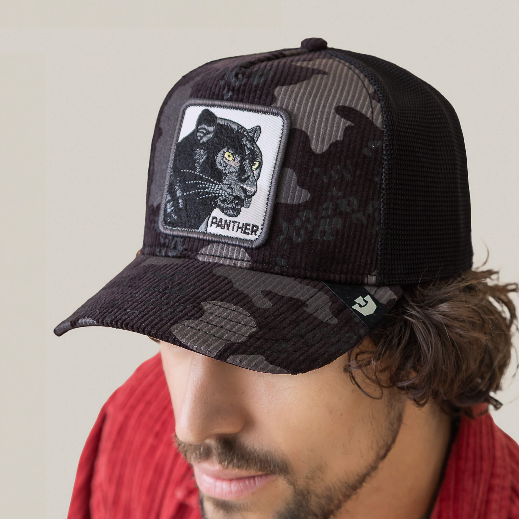 A man wearing a Goorin Bros. Panther Camo cap, which features black stealth camo mesh and a front patch with a panther illustration and "PANTHER" text, paired with a red shirt.