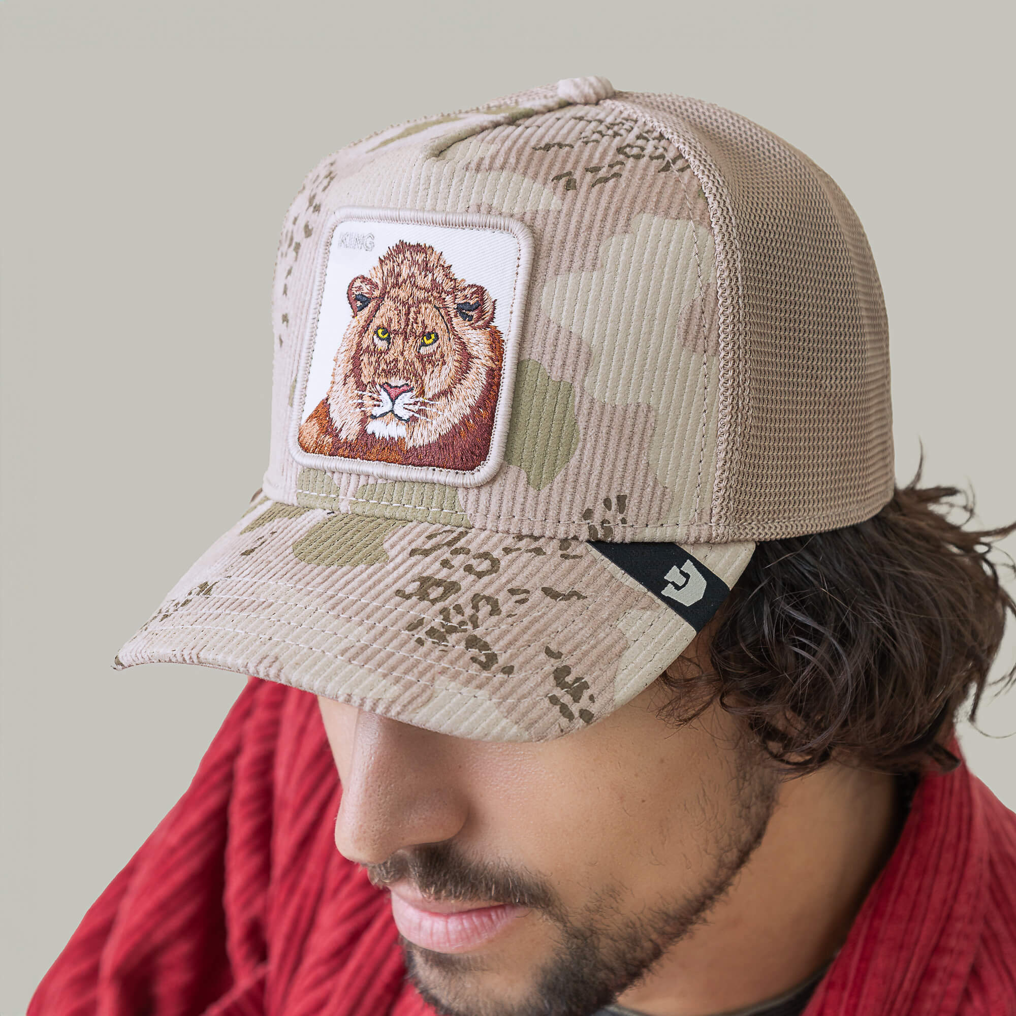 A man in a red shirt wears the Goorin Bros. King Camo beige camouflage mesh cap with a lion face patch, pictured against a plain background.