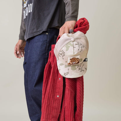 A person in dark jeans and a long-sleeve shirt holds a red corduroy jacket and the King Camo hat with tiger patch by Goorin Bros. in their left hand.