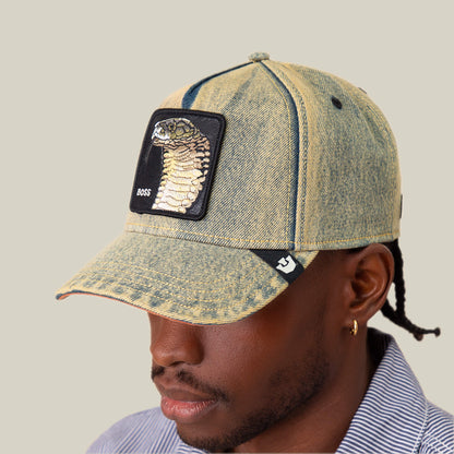 Someone wears Goorin Bros.’ Pit Boss denim hat featuring a striking Cobra patch and the word BOSS, styled with a striped shirt and a small hoop earring.