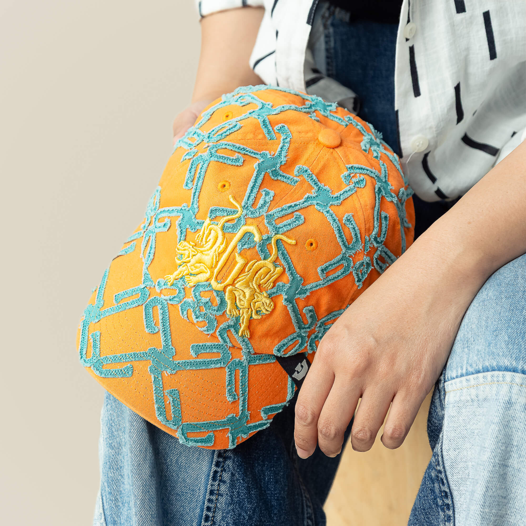 A person sits in blue jeans and a striped white shirt, holding the Goorin Bros. 'Future Baby Daddy' cap—orange with blue chain embroidery and a yellow lion emblem—radiating main character energy.