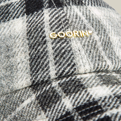 Close-up of the Goorin Bros. Bespoke Wordmark cap, a black, white, and gray plaid wool blend design with a gold Goorin metal logo near the seam.