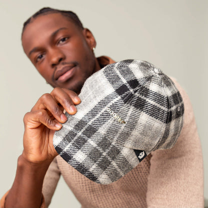A man holds out the Goorin Bros. Bespoke Wordmark cap, a gray and black plaid wool blend hat, with the logo on the side, partially obscuring his face.