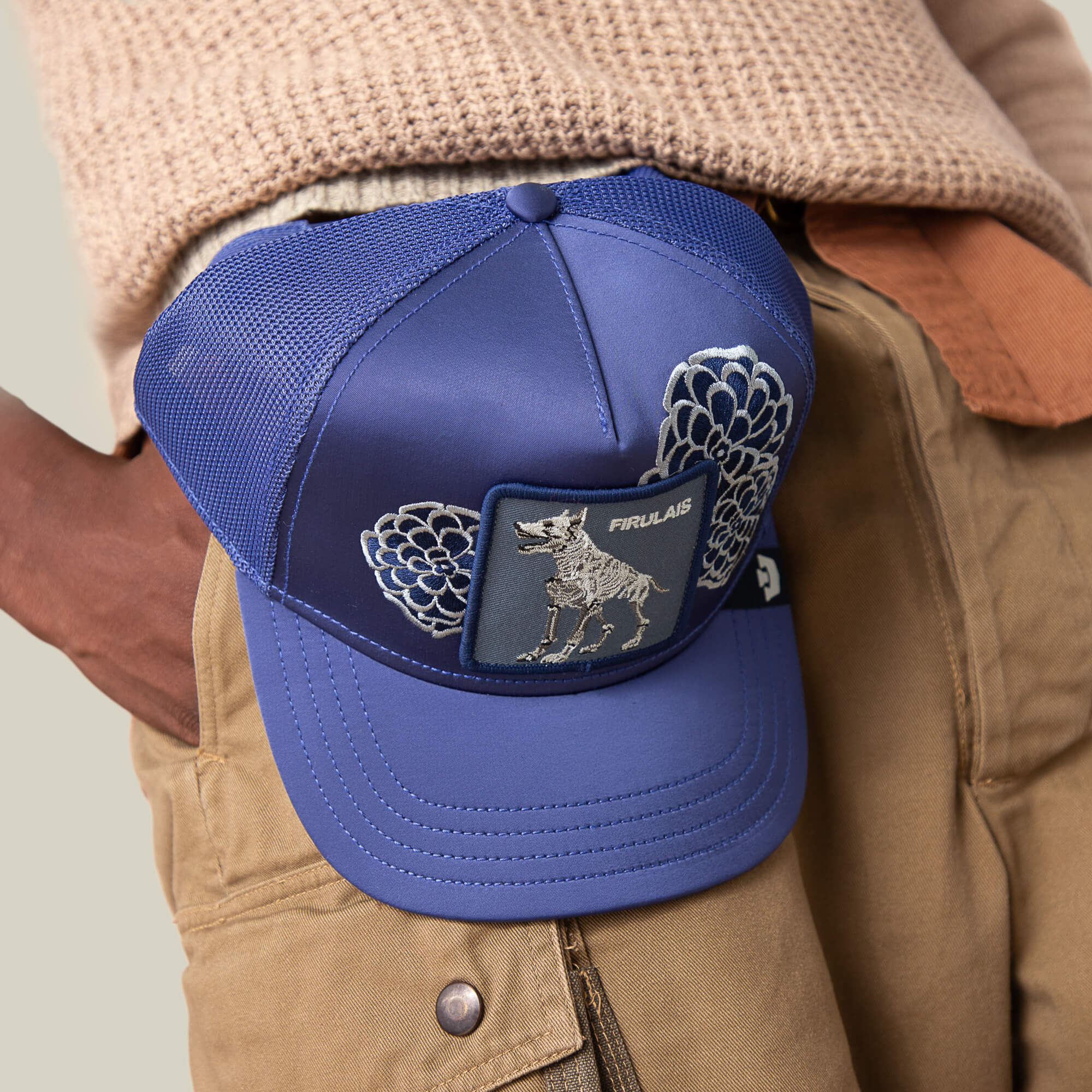 Someone wearing brown pants and a beige sweater holds the Goorin Bros. Dia De Firulais blue mesh baseball cap featuring an animal skeleton patch and unique designs—perfect for Life After Ordinary.