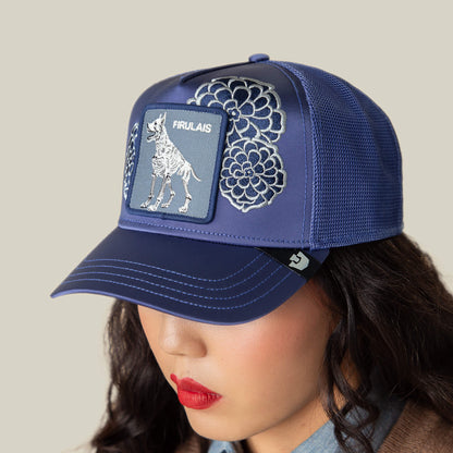 Person wears a blue mesh baseball cap by Goorin Bros., called Dia De Firulais, featuring embroidered floral patterns and a Firulais patch, celebrating Life After Ordinary.