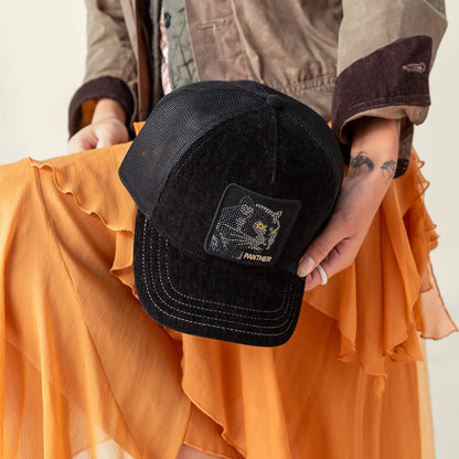 Person holding the Goorin Bros. Iced Panther black mesh trucker hat with a luxury panther patch, styled with an orange pleated skirt and brown jacket.
